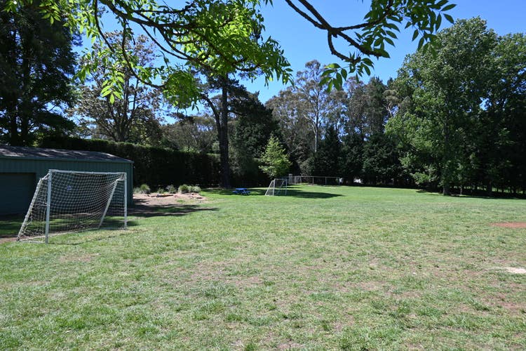 A grassy school sports field with two soccer goals, surrounded by tall trees and a storage shed to the left under clear blue skies.
