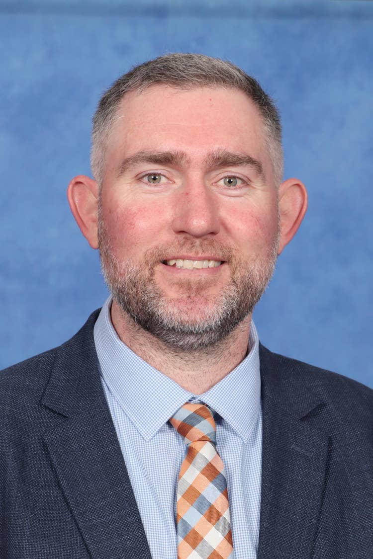 Smiling man with short brown hair and a beard, wearing a suit jacket and light blue shirt, in front of a blue background.
