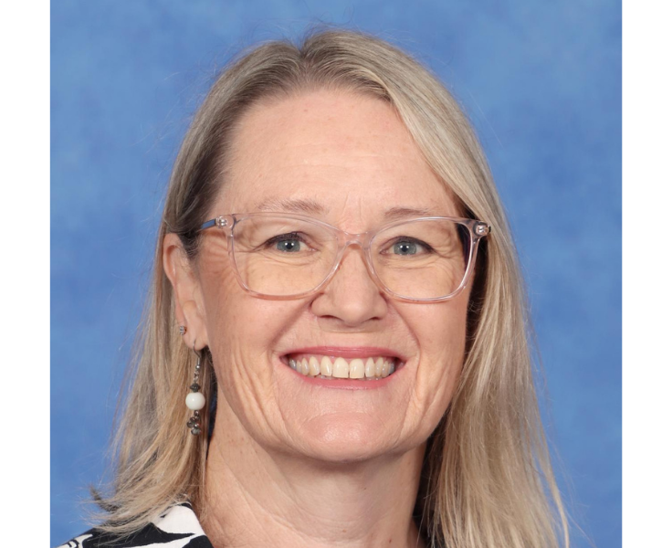 Smiling woman with blonde hair and clear glasses, wearing drop earrings and a patterned top, in front of a blue background.