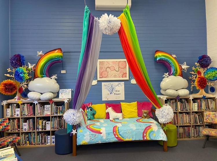 Colourful bed between two shelves of library books. Colourful rainbow canopy above the bed, rainbow and unicorn decorations to either side.