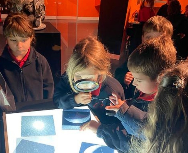 Young students in school uniforms explore a science exhibit; one child uses a magnifying glass to examine illuminated images, while others observe with curiosity.