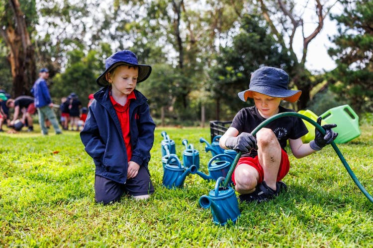 Two young students outdoors; one fills a blue watering can using a green hose while the other watches. More watering cans are lined up on the grass.