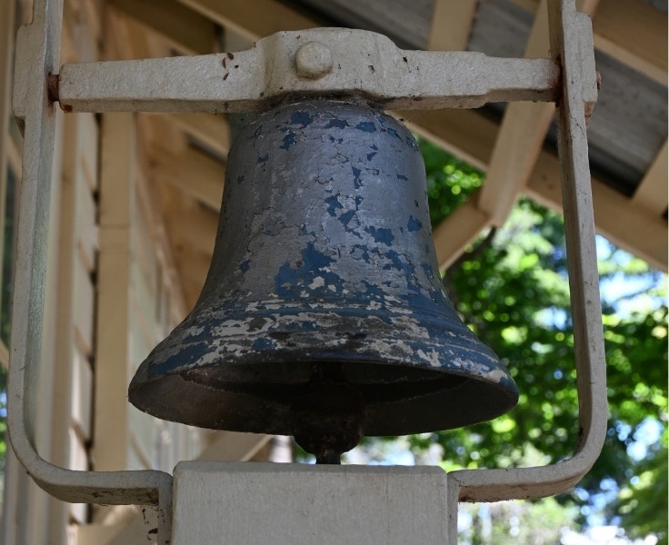 Close-up of an old, weathered school bell mounted on a metal frame outside a building with wooden siding and a corrugated roof.