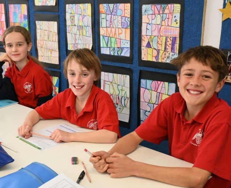 Three primary school students in red uniforms sit at a classroom table, smiling while working on an activity. Colorful art is displayed on the wall behind them.