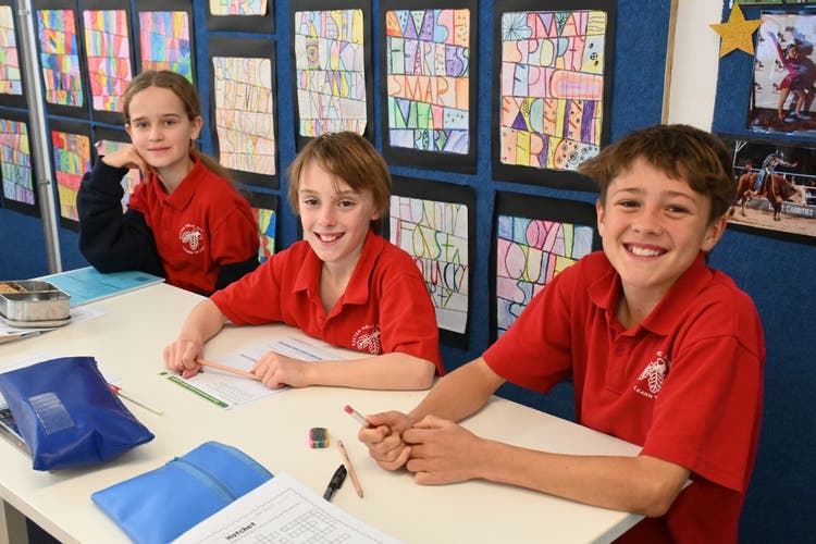 Three students in red school uniforms sit at a desk in a classroom, smiling while working on an activity. Colorful artwork is displayed on the wall behind them.