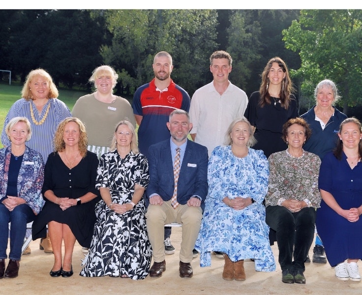 Group photo of school staff members posed outdoors in two rows, with trees and a sports field in the background. The group includes both men and women dressed in professional and casual attire, smiling at the camera.