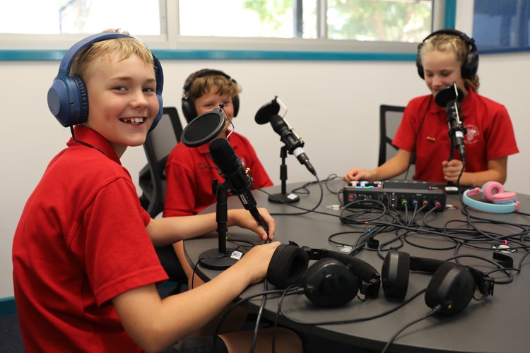 Three primary school students wearing headphones and using microphones in a classroom podcast studio, smiling and engaged.