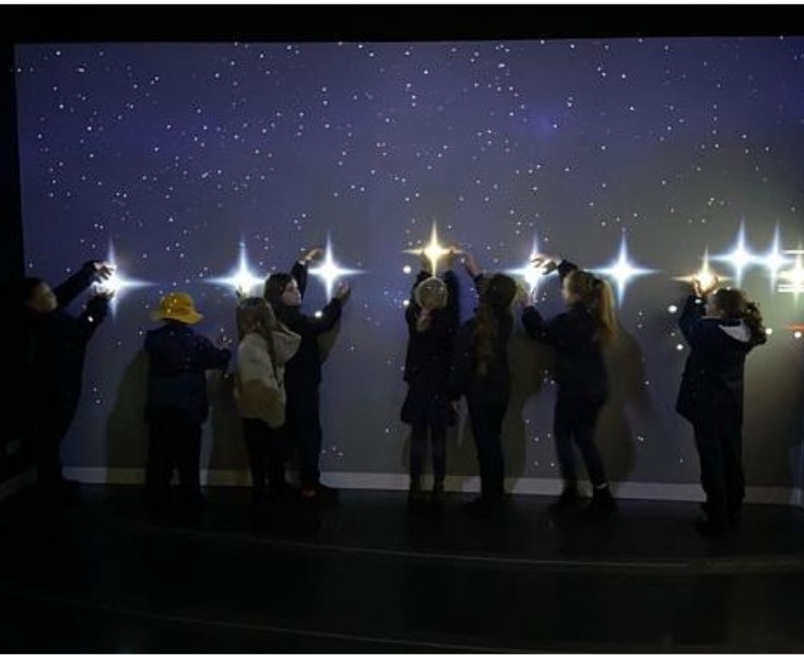 A group of children stands in front of a dark, starry wall display, reaching out to touch bright interactive lights shaped like stars. The scene is softly lit, creating a magical, immersive atmosphere.
